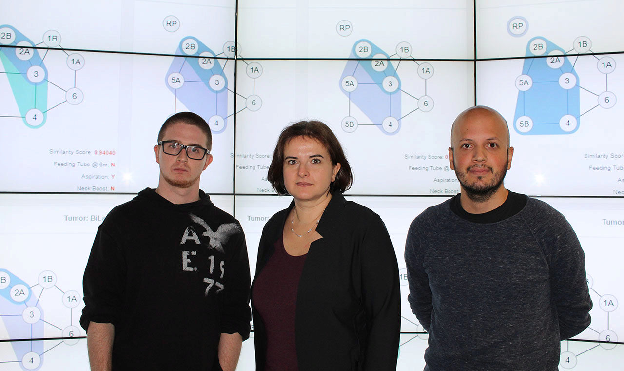 Liz Marai with graduate students Tim Luciani and Juan Trelles Trabucco standing in EVL&rsquo;s CAVE2 hybrid reality system with diagrams of patient data depicting head and neck lymph nodes affected by disease spread.